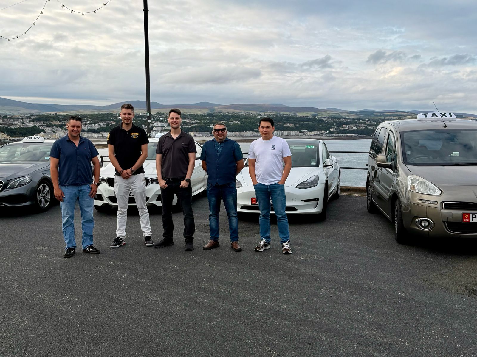 One Island Taxi team and participating local taxi drivers with vehicles overlooking Douglas Bay, Isle of Man